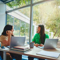 Two gray HP laptops open on a bright office table with notebooks and coffee mugs, with two women conversingNearby.