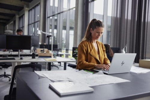 Silver HP EliteBook laptop open on desk with woman typing in modern office setting.