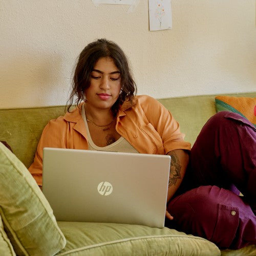 Silver HP laptop open on a green couch with a person wearing an orange shirt and maroon pants using it.
