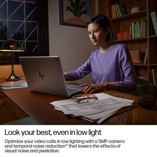 Slim silver HP EliteBook 14-inch laptop open on wooden desk with papers and glasses, woman typing.
