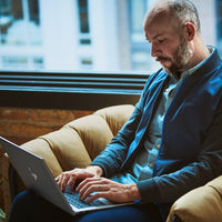 Man in blue blazer using a grey HP ZBook laptop on a tan sofa by a large window.
